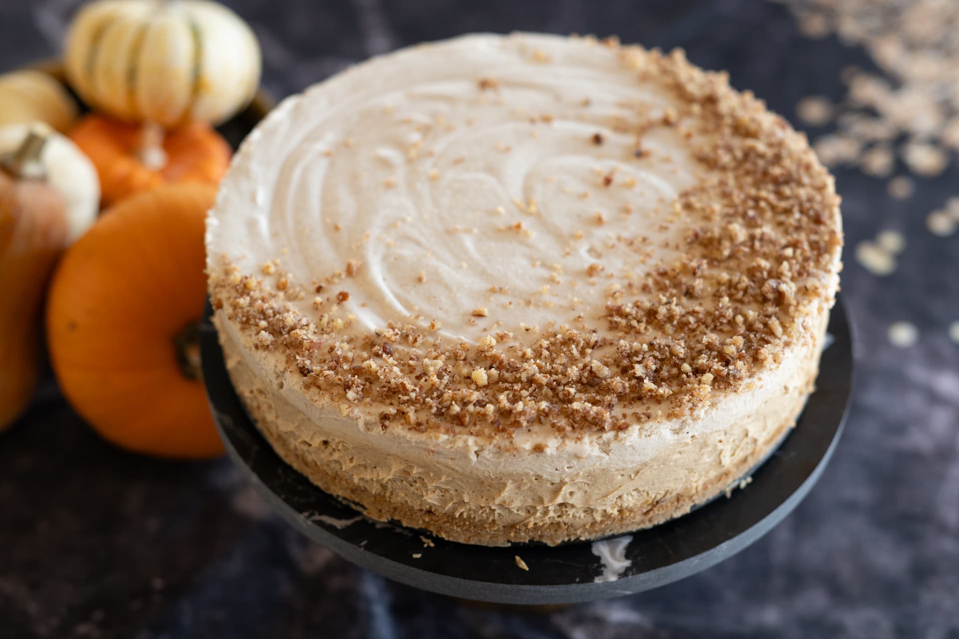 A pumpkin-spiced cake with swirled frosting and a graham crumble, surrounded by mini pumpkins