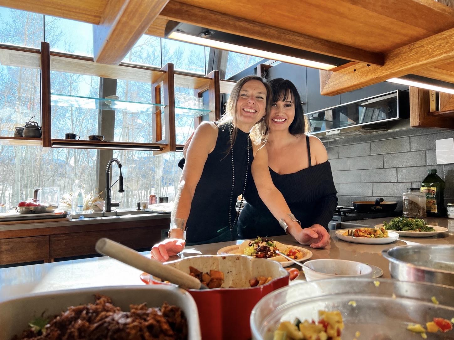 Two women smiling together in a sunlit kitchen behind a spread of shared dishes