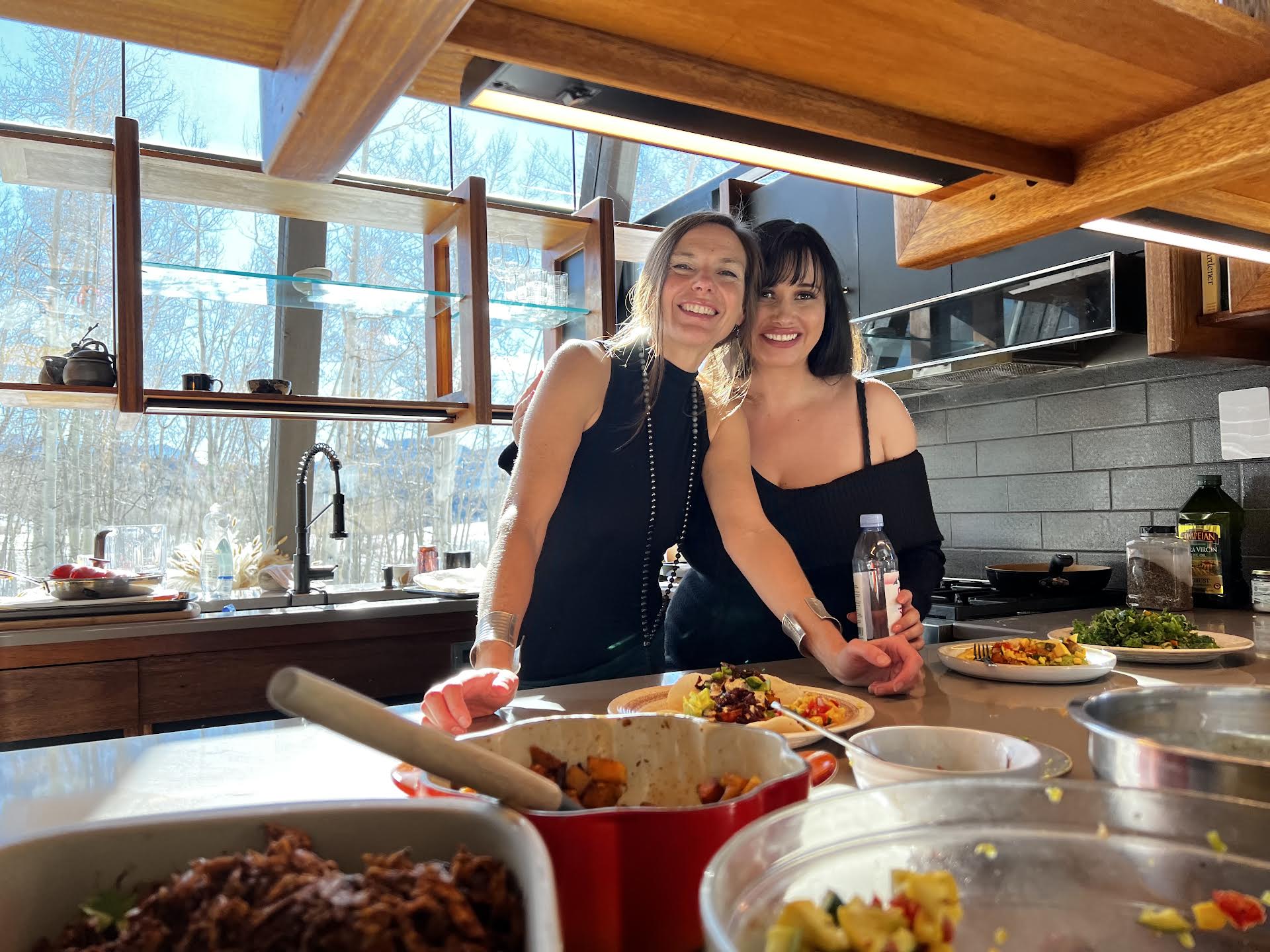 Caitlin and a guest laughing behind a family-style spread in a sunlit kitchen